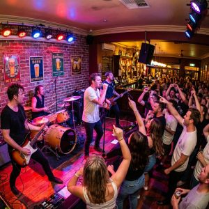 An energetic live band performing on stage at an Australian pub with warm stage lighting and a packed crowd